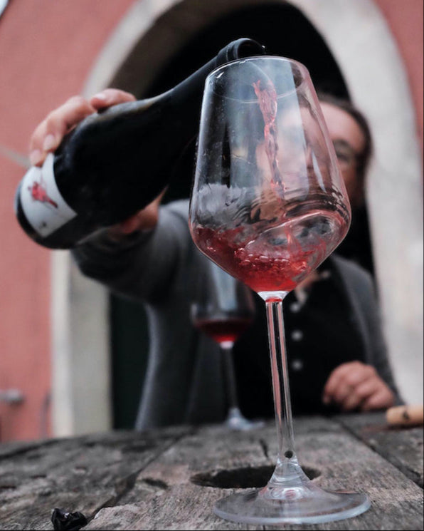 Closeup picture of a glass of wine in the forefront, with wine being poured from a bottle held by a person in the background.