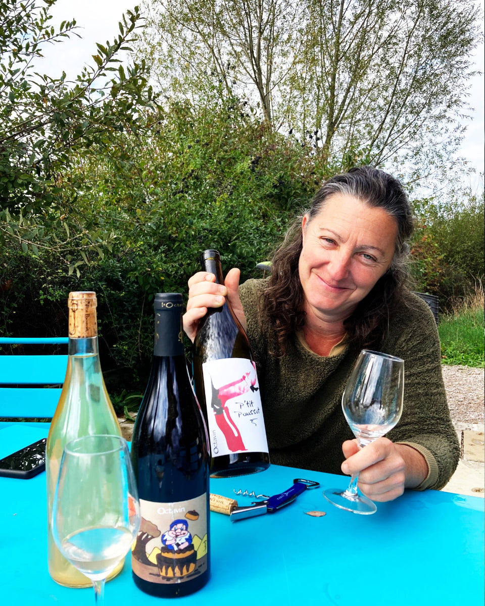 Picture of Alice Bouvot, owner of L'Octavin, sitting at a table and showing three bottles of her wines.